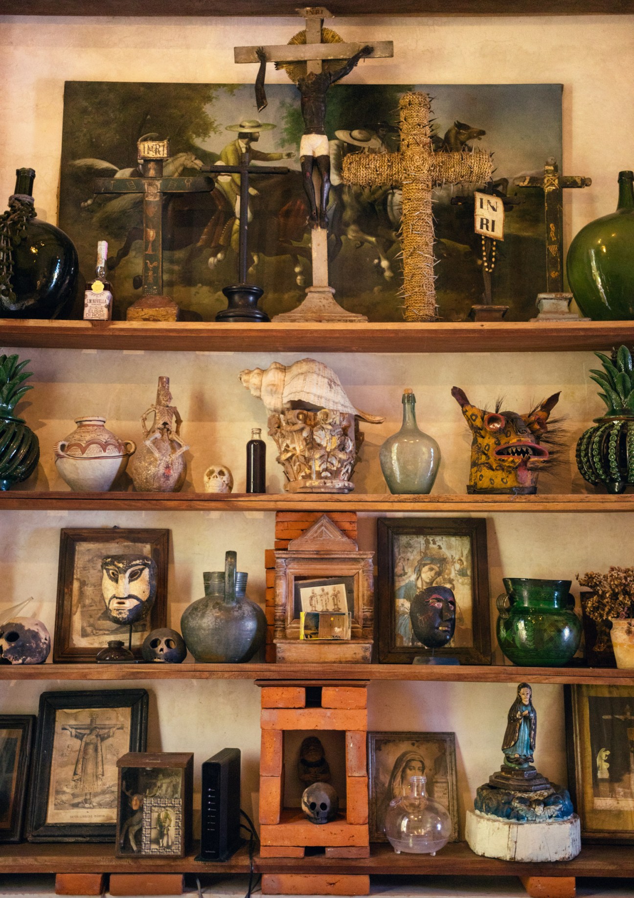 Wooden shelves displaying eclectic religious artwork, artifacts, ceramics, skulls and decorative objects including crucifixes and framed prints.