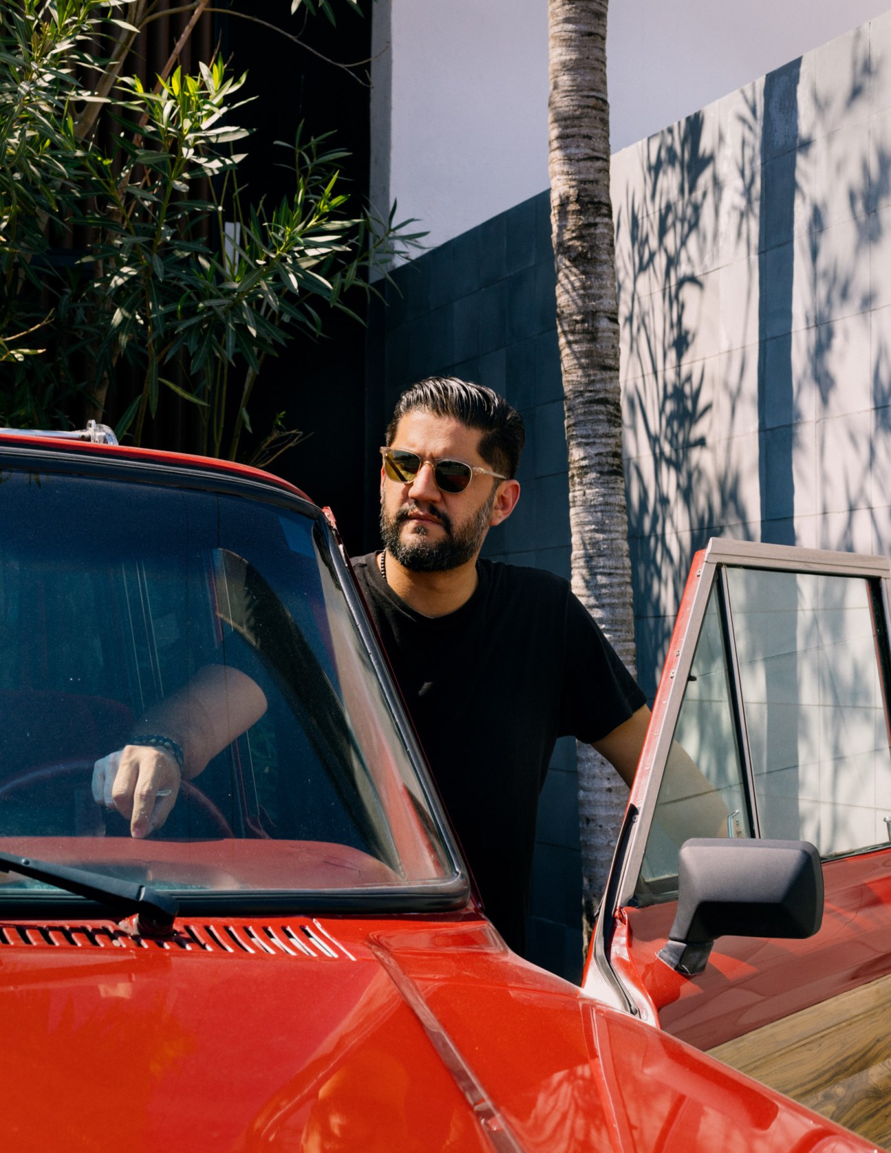 Carlos Martínez standing beside a red 1980s Jeep wearing sunglasses and a black shirt.