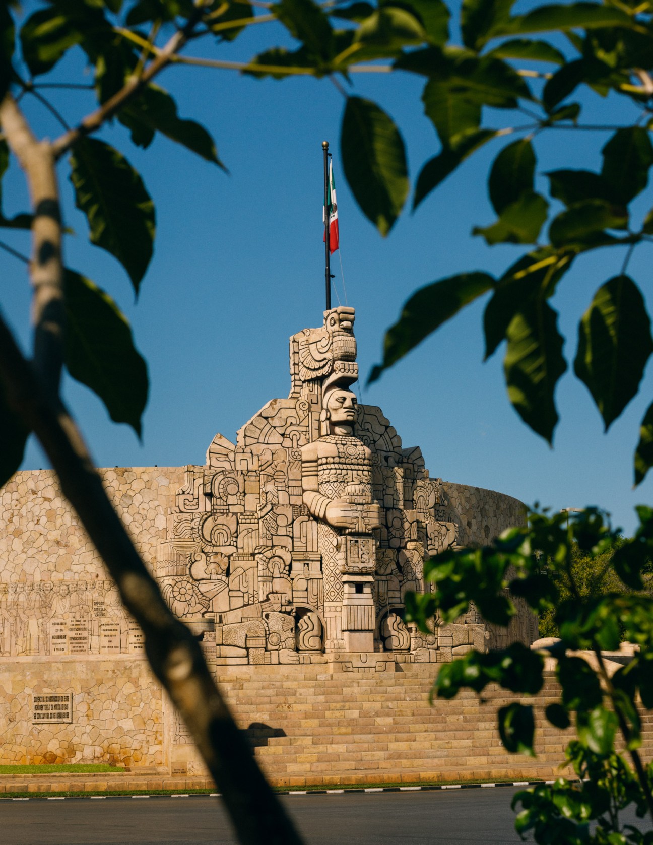 Monumento a la Patria monument with Mayan-inspired carved stone sculpture and Mexican flag under blue sky with tree branches in foreground.