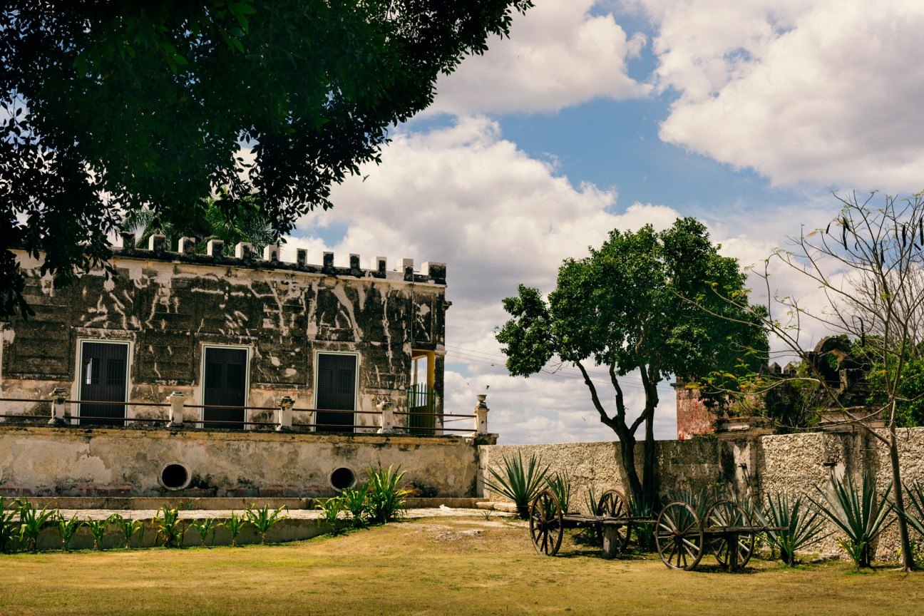 Weathered colonial hacienda building with stone facade, arched windows, and manicured grounds under blue sky.
