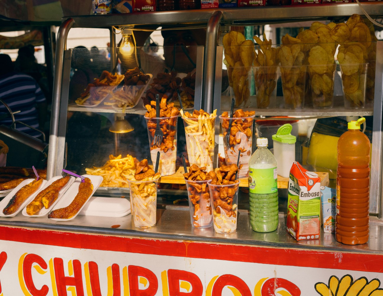 Food stand display with fried snacks in cups, sausages on plates, and beverages in Plaza Grande, Mérida.