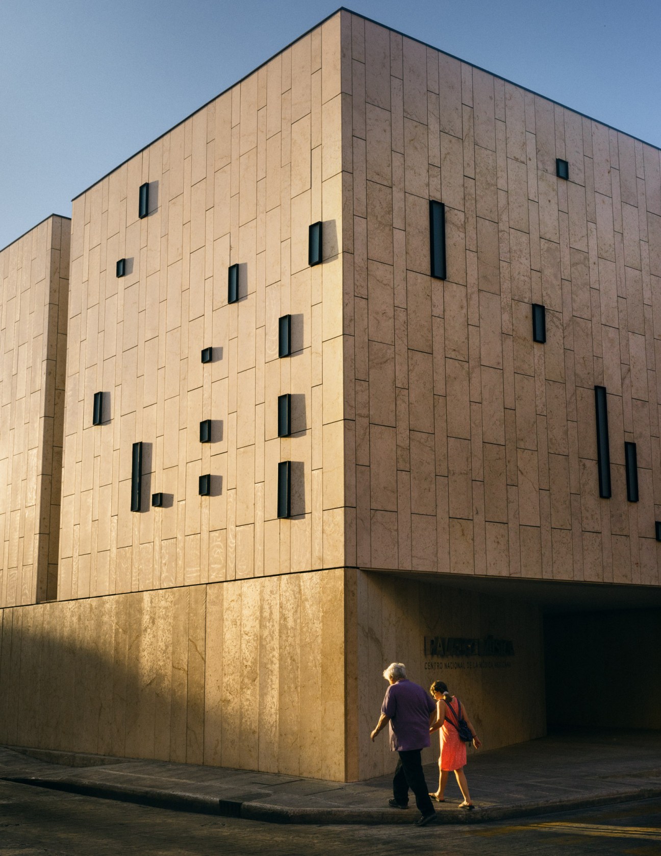 Modern beige and brown stone building with narrow window openings and two pedestrians walking past its base.