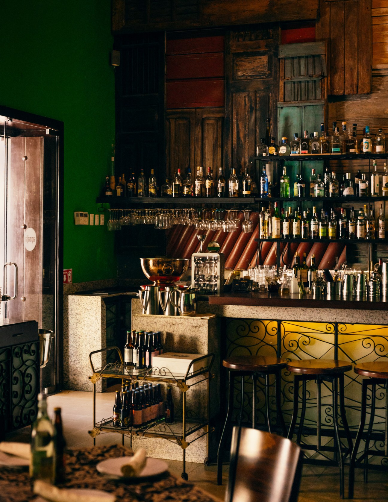 Interior of Apoala, an Oaxacan restaurant in Mérida, with green walls, wooden doors, and shelves of spirits behind the bar.