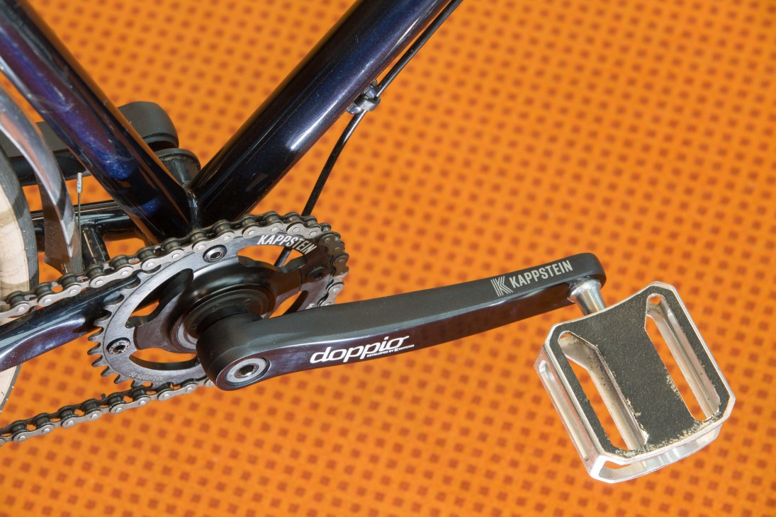 Close-up of Kappstein bike components showing chainring and crank arm with orange textured background.