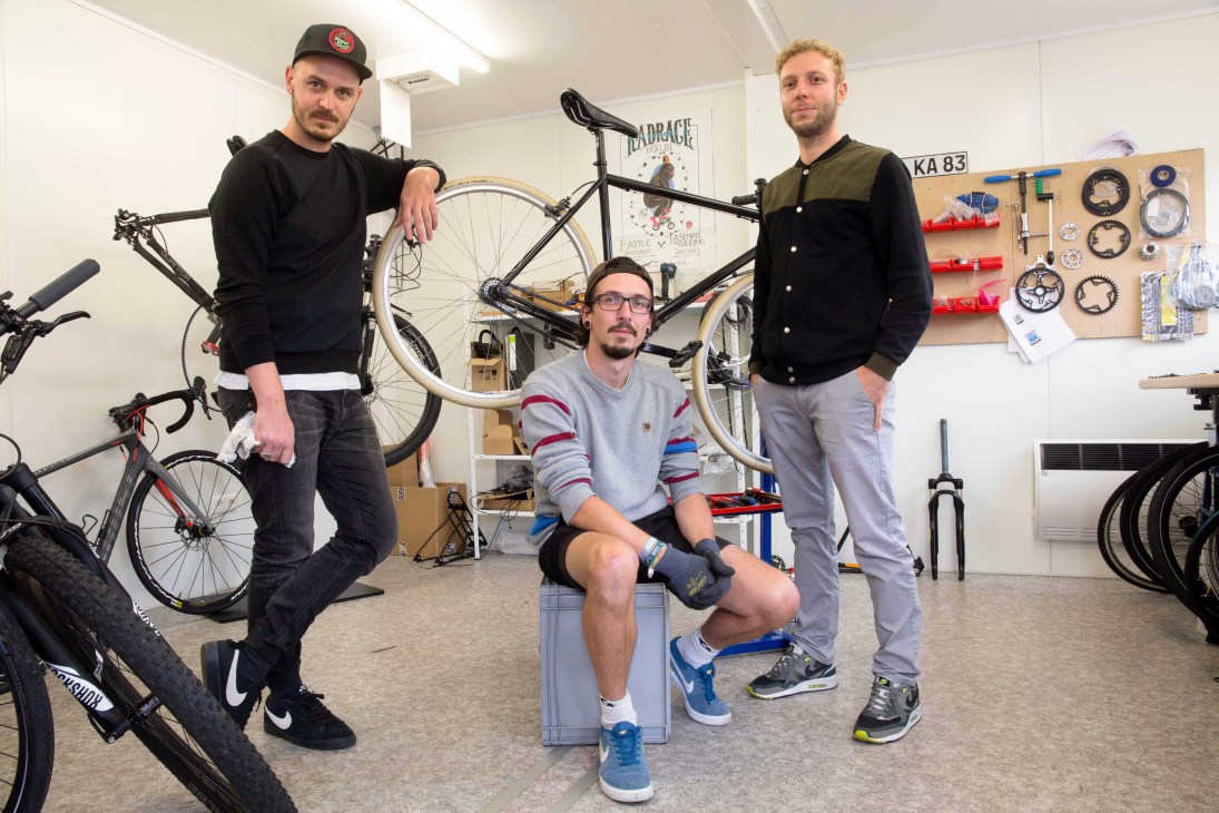 Three men posing in a bike assembly workshop with tools and components on the wall behind them.