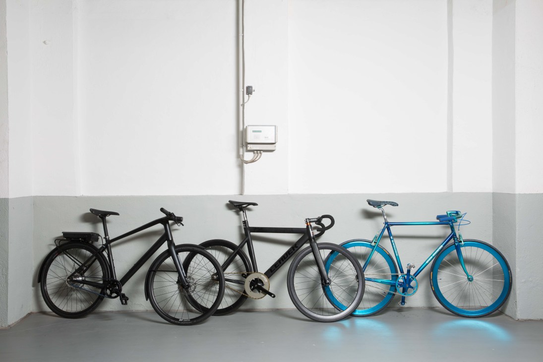Four bikes leaning against a white wall with a charging station mounted above.