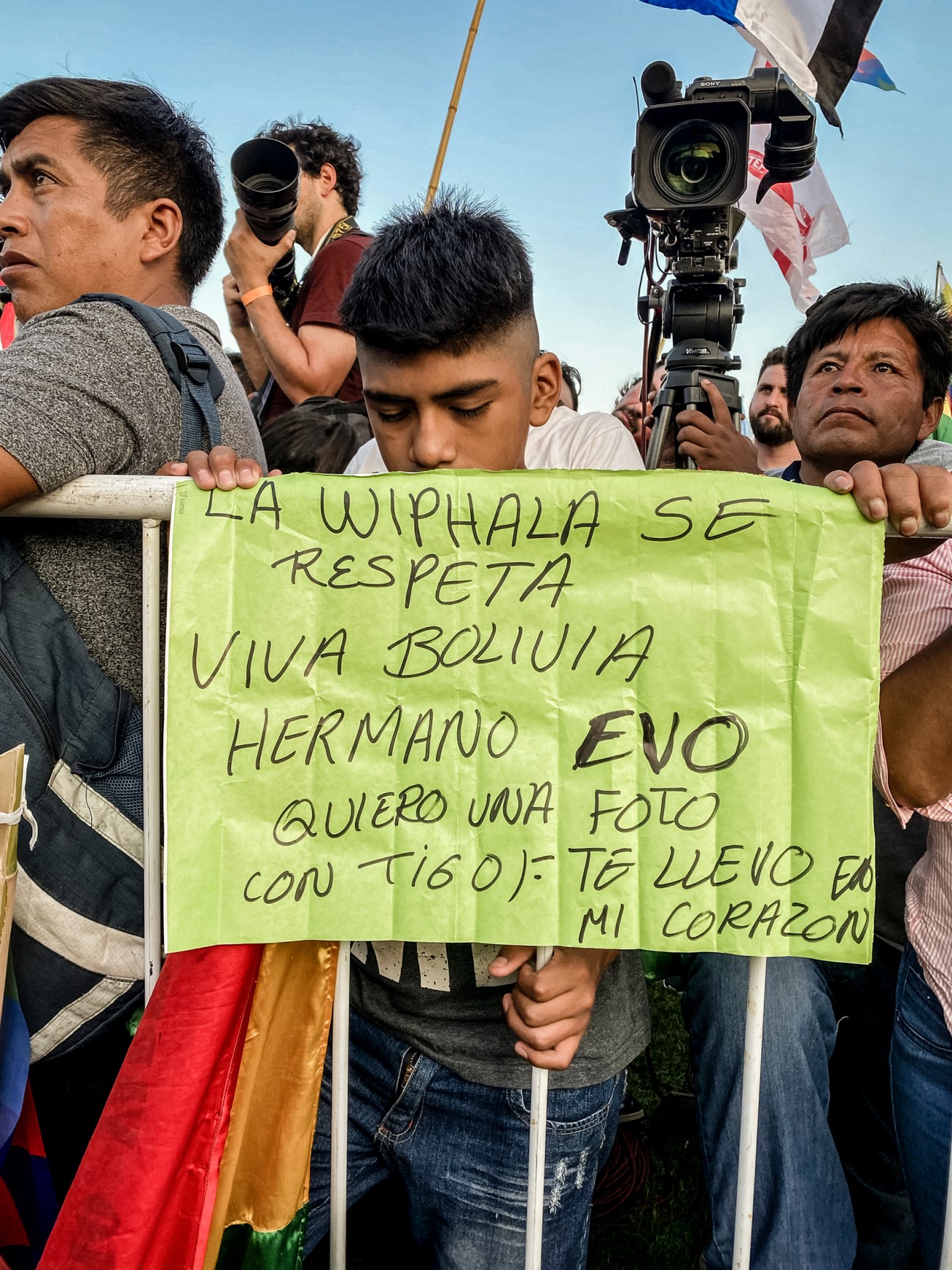 Young man holding green sign in Spanish reading 'LA WIPHALA SE RESPETA VIVA BOLIVIA HERMANO EVO' at protest