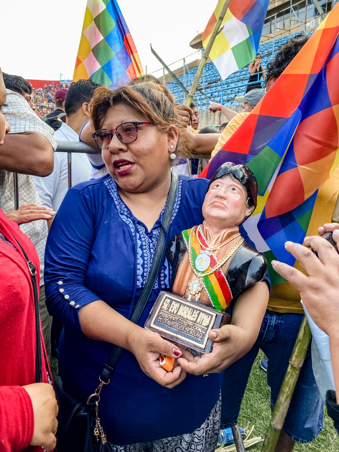 Woman in blue holding small figurine of Evo Morales at political rally with colorful flags