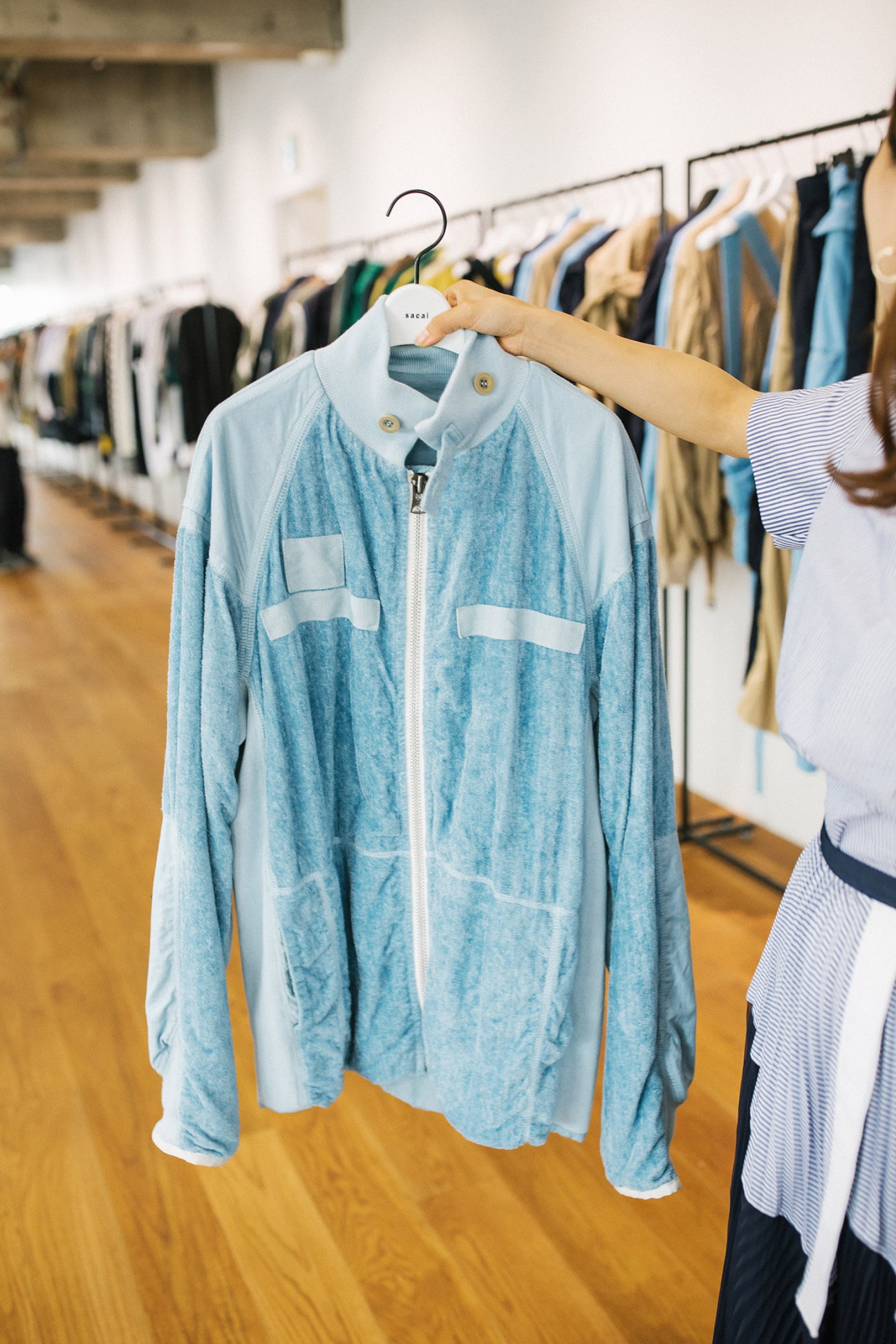 A hand holds a Sacai jacket on a hanger in the studio, showing the brand's signature mixed-fabric design with denim and white cotton panels.