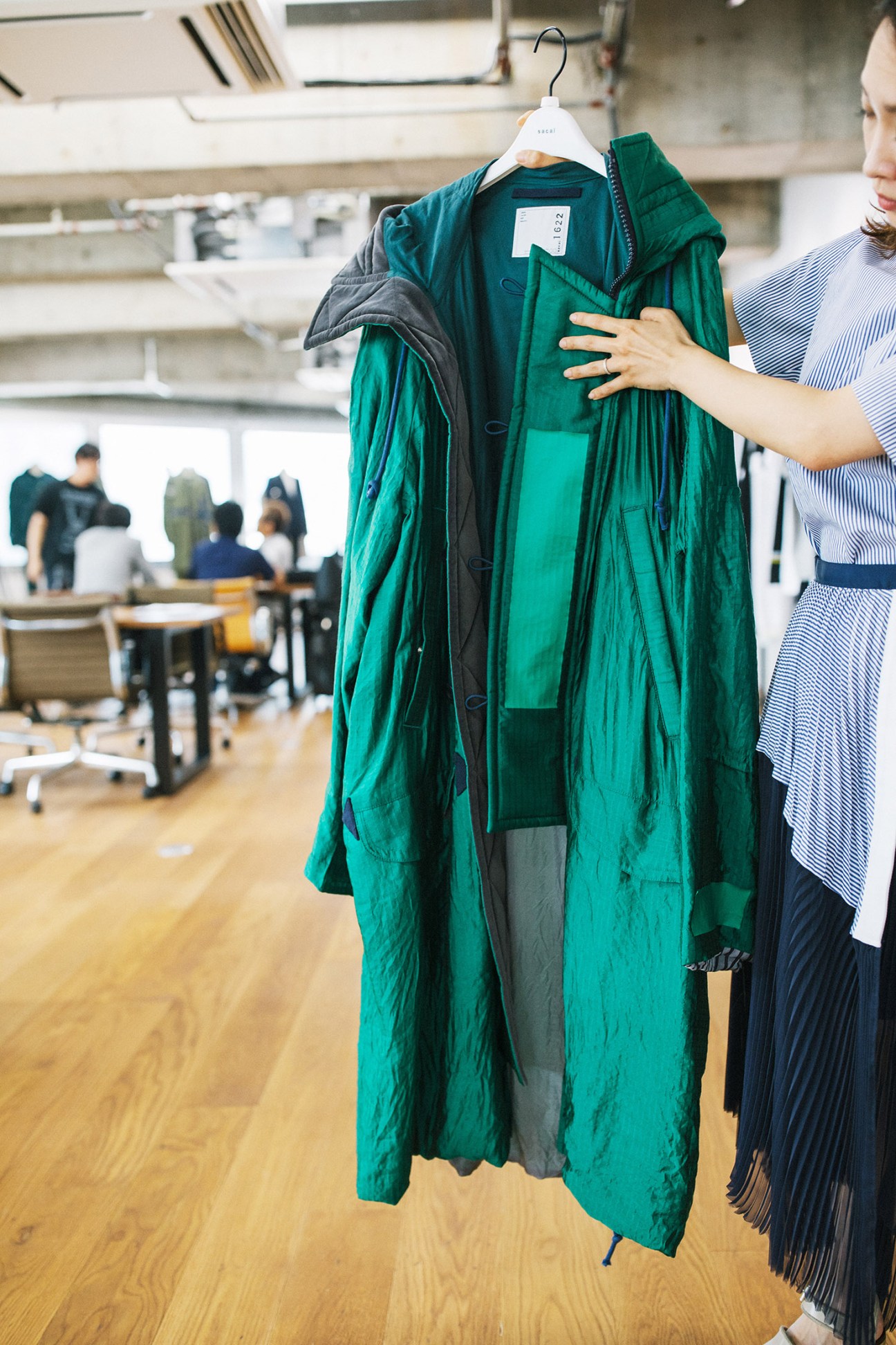 A woman holds a green and black layered Sacai jacket on a hanger in the studio.