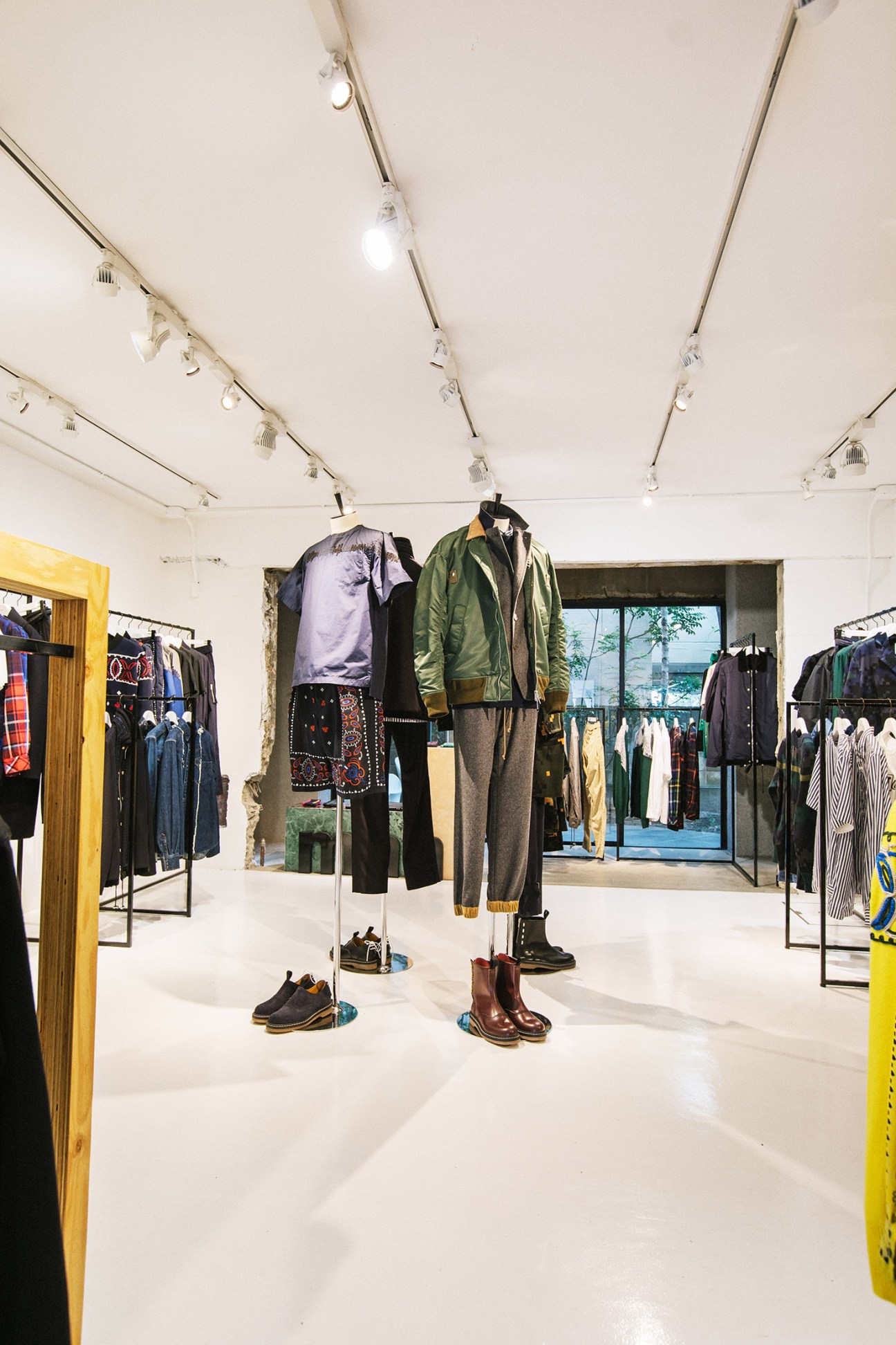 Interior of Sacai flagship store with minimalist white space, clothing displays on racks, and mixed material textures.