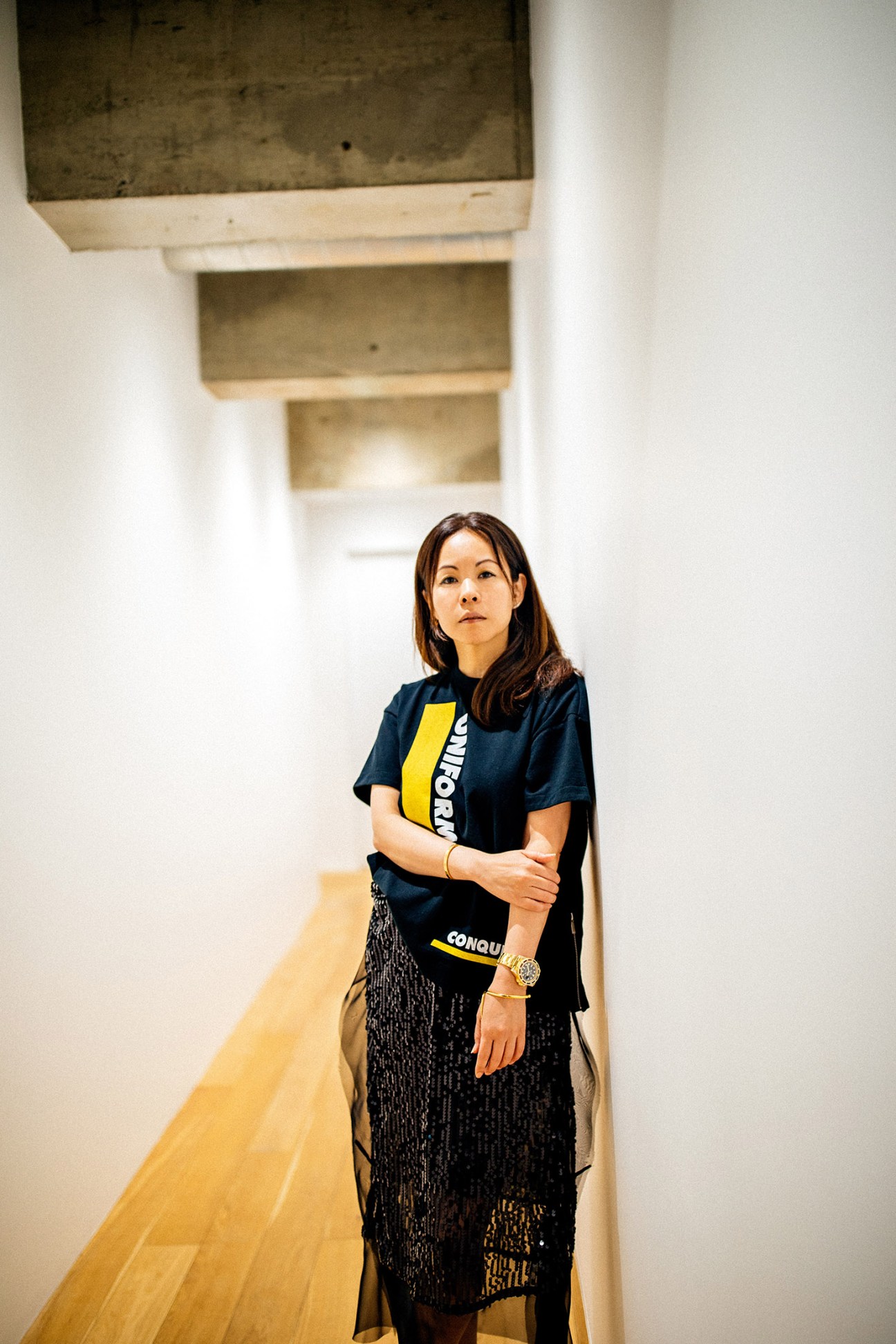 Chitose Abe stands in a minimalist corridor wearing a navy t-shirt with yellow and white graphic design paired with a patterned skirt.