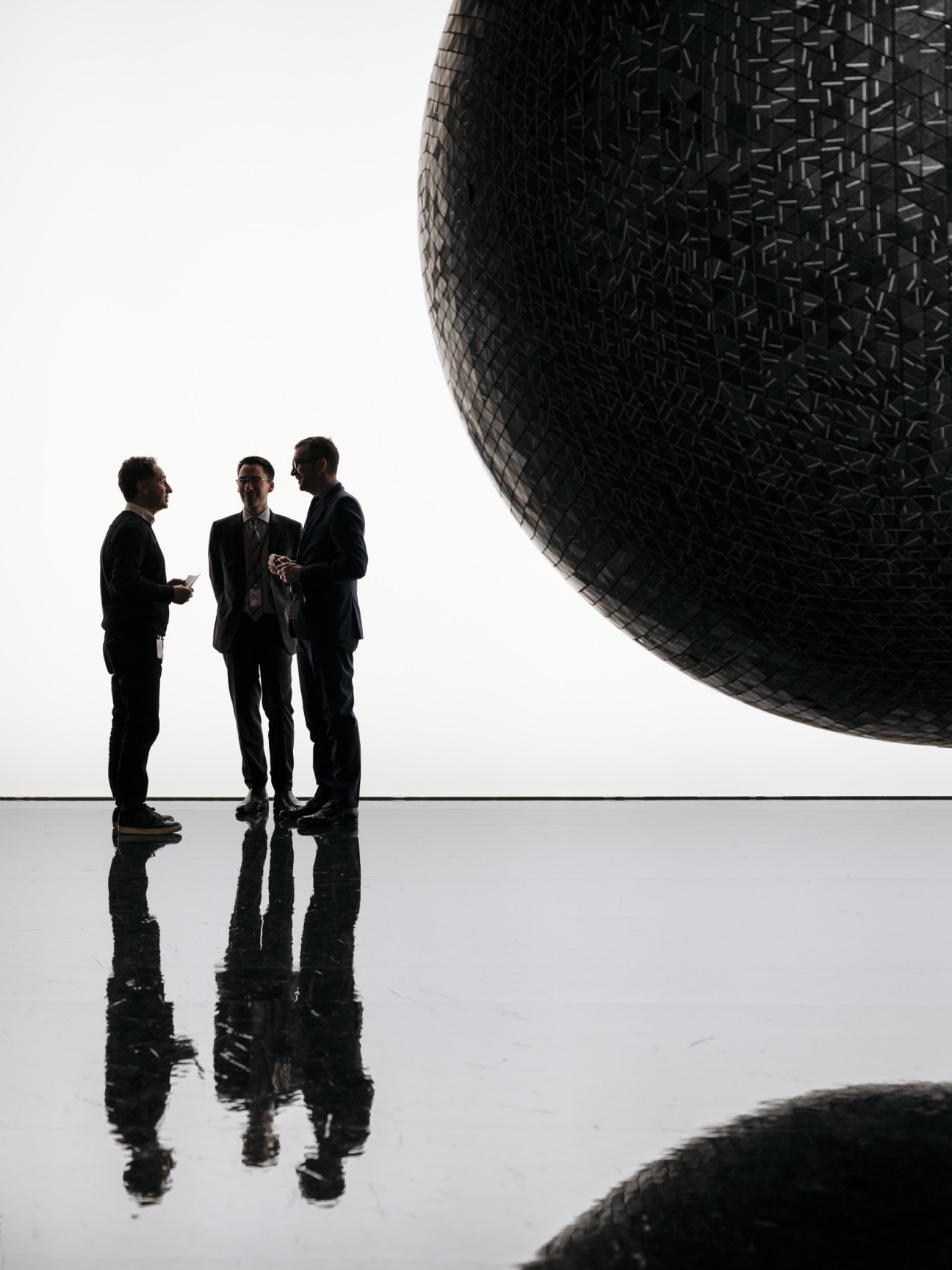 Three men in dark business suits conversing beneath a large spherical black sculptural installation.