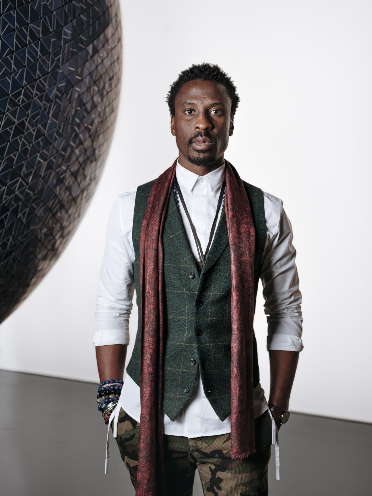 Man wearing a green plaid vest, white shirt, and burgundy scarf posed against a geometric patterned wall.
