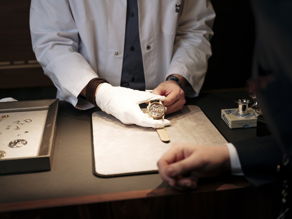 Watch craftsperson in white shirt and gloves displaying a luxury watch at a workbench.