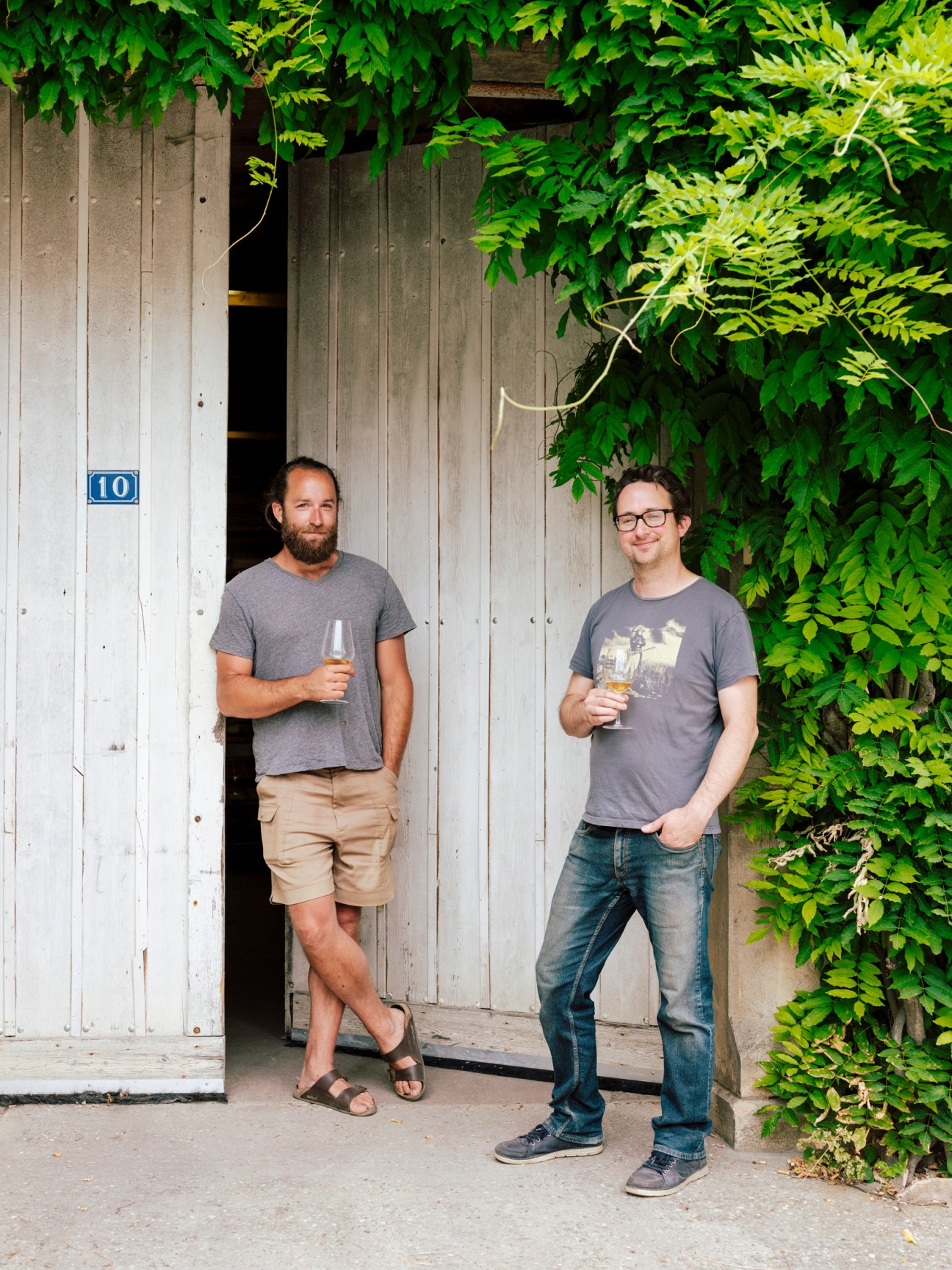 Two men holding wine glasses stand outside a weathered wooden door covered in ivy vines.