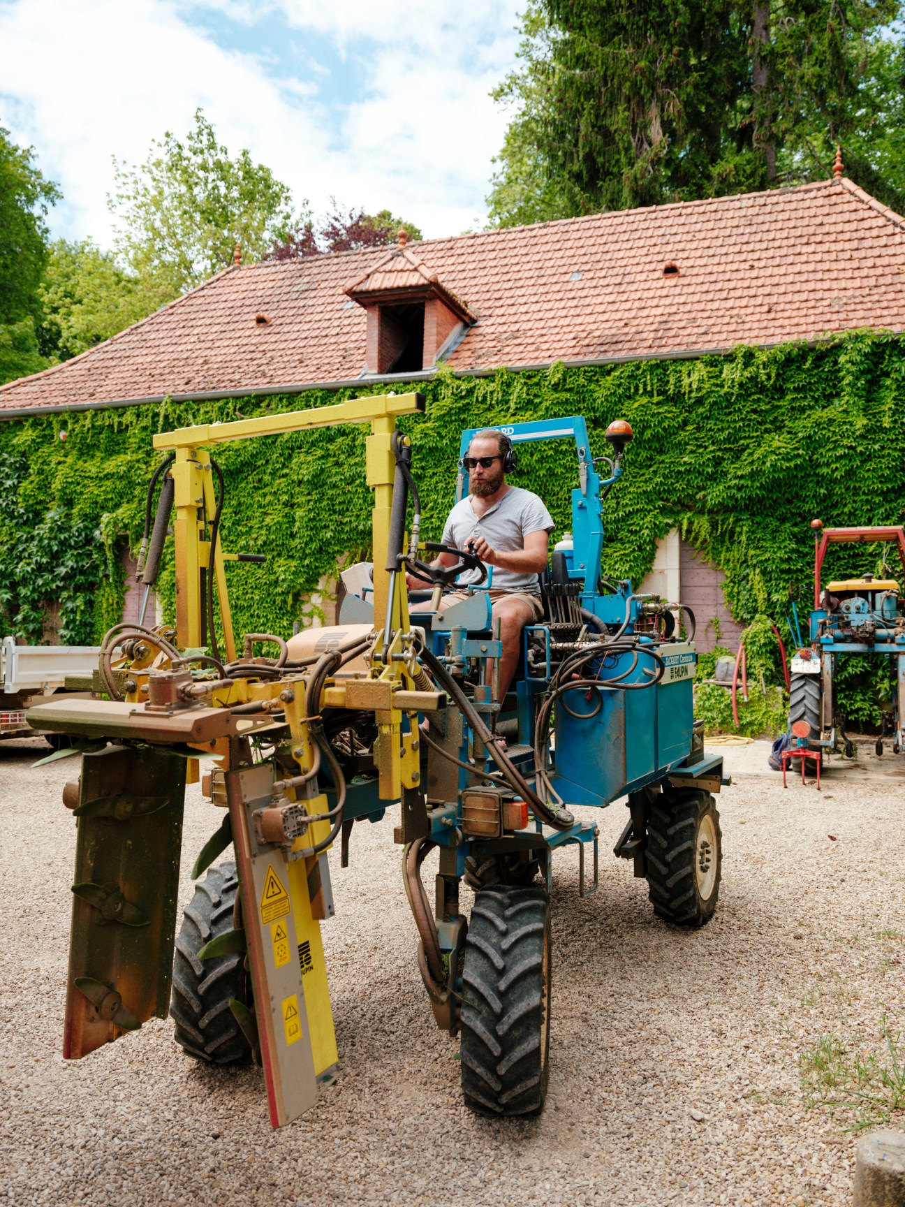 Jon Purcell sits atop a yellow tractor in a vineyard courtyard surrounded by ivy-covered buildings.
