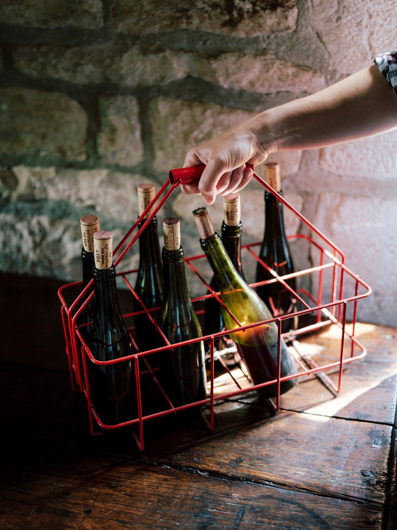 Hand holding a red metal wine bottle carrier with multiple wine bottles against a stone wall.
