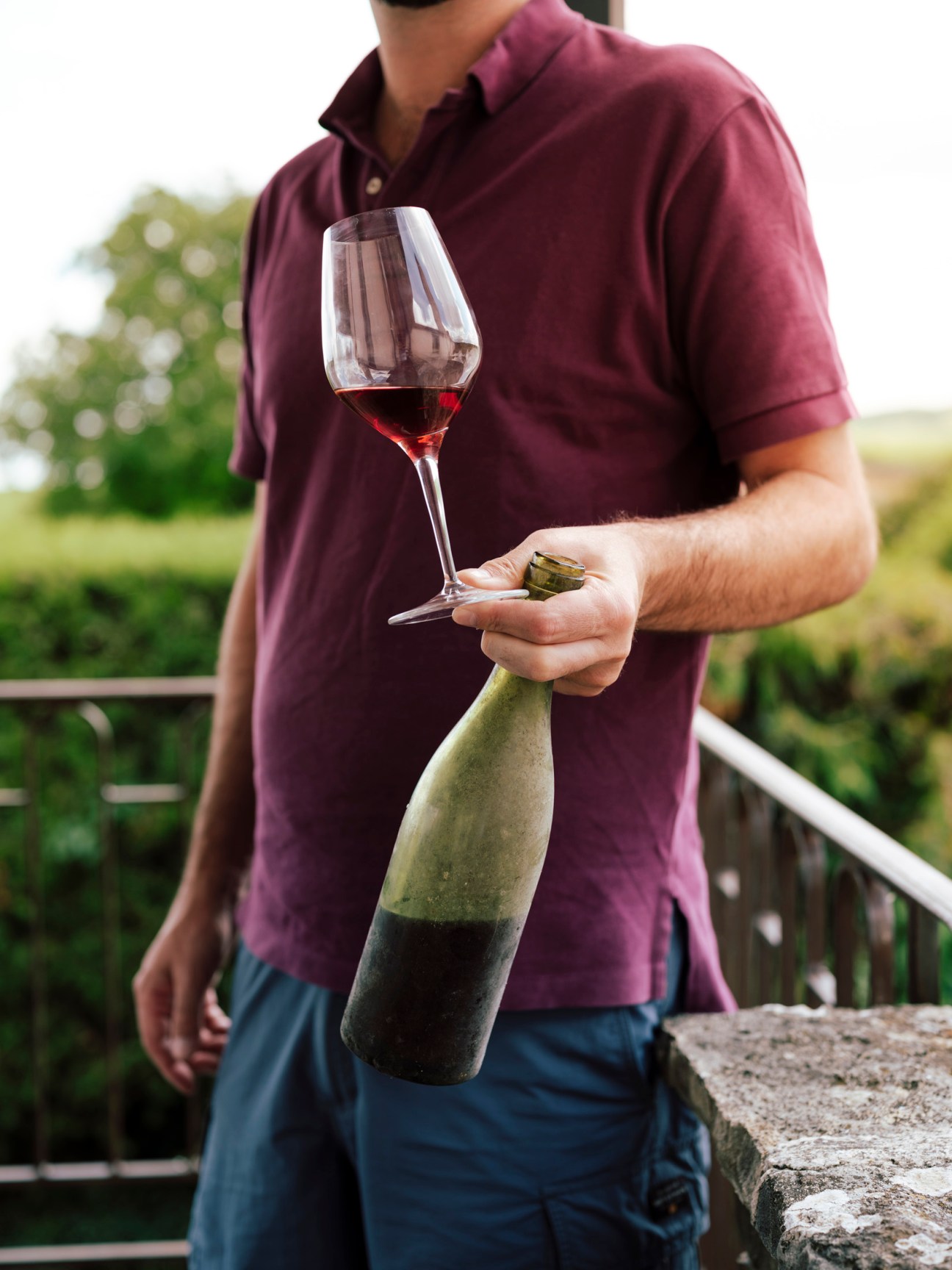 Man in burgundy shirt holding a wine glass and bottle in a vineyard setting.