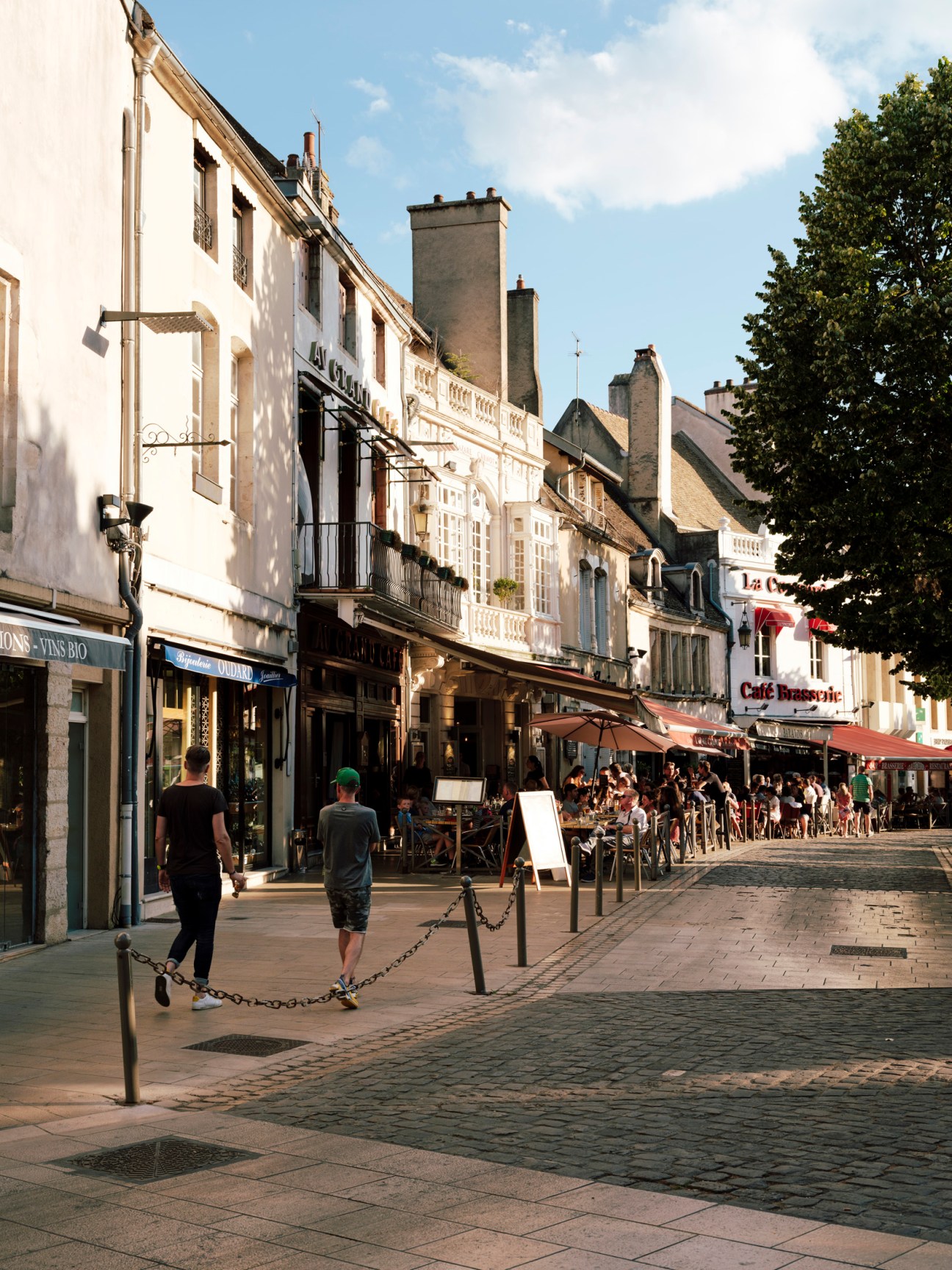 Pedestrians stroll along a sun-lit street lined with historic buildings, shops and cafés in Beaune, France.