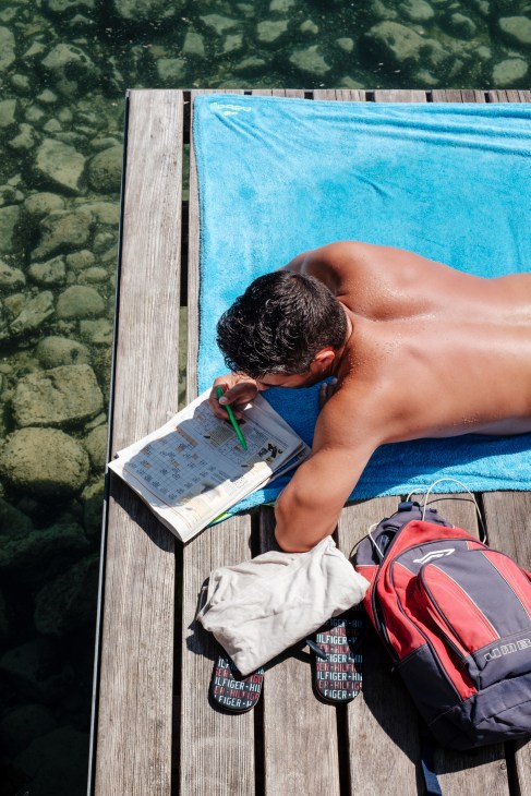 Person solving a Sudoku puzzle while sunbathing on a blue towel at Bains des Pâquis jetty.