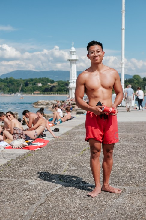 Young man in red swimming shorts standing on the beach at Bains des Pâquis with Lake Geneva and mountains in the background.