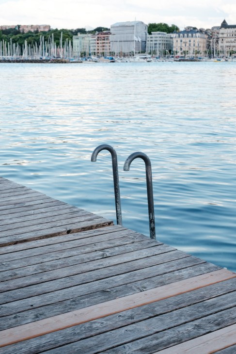 Metal swimming ladder on wooden dock beside Lake Geneva with Geneva's old town and sailboats visible across the water.