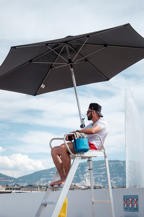 Lifeguard sits on elevated chair under black umbrella overlooking Lake Geneva at Bains des Pâquis.
