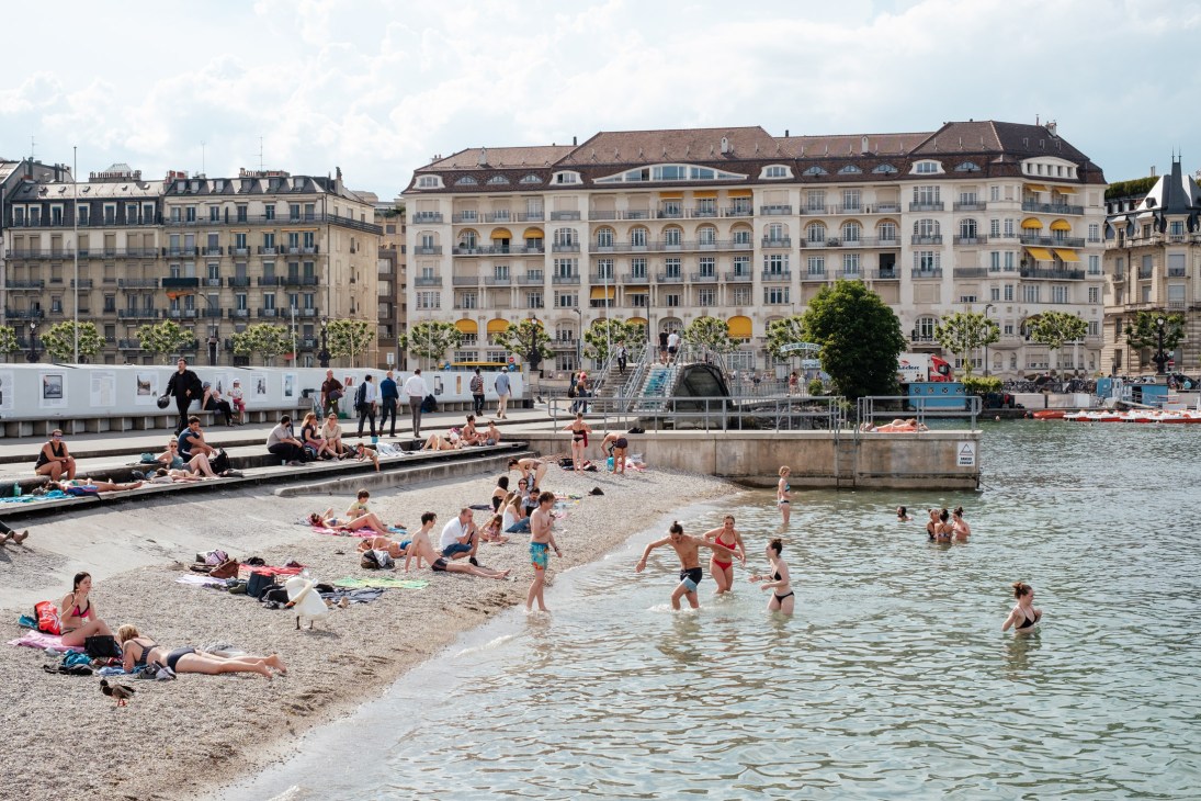 Sunbathers and swimmers enjoy Lake Geneva's beach at Bains des Pâquis with historic buildings behind.