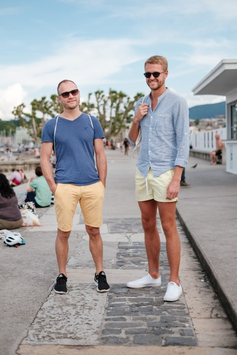 Two men standing on a jetty at Bains des Pâquis with Lake Geneva and the old town visible in the background.