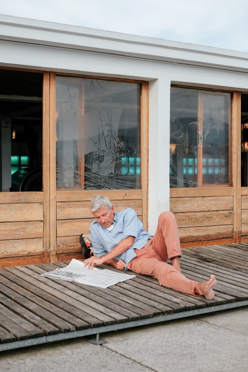 Older man in blue shirt and pink pants doing Sudoku on wooden deck at Bains des Pâquis.