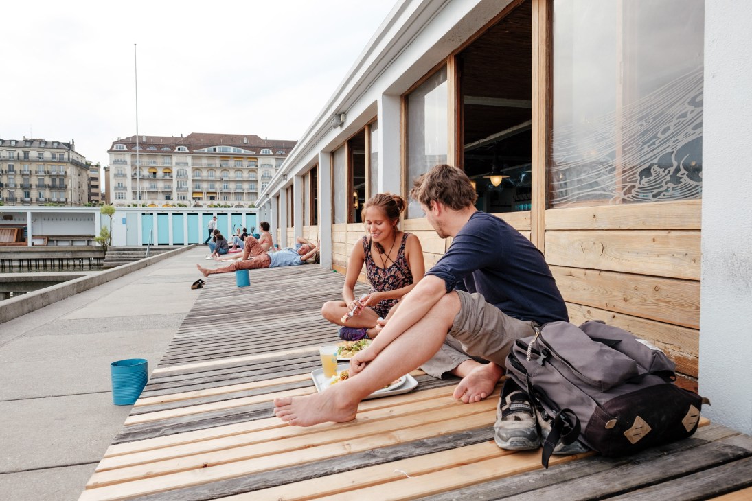 Two visitors enjoy lunch while seated on a wooden deck at Bains des Pâquis with Geneva's old town visible in background.