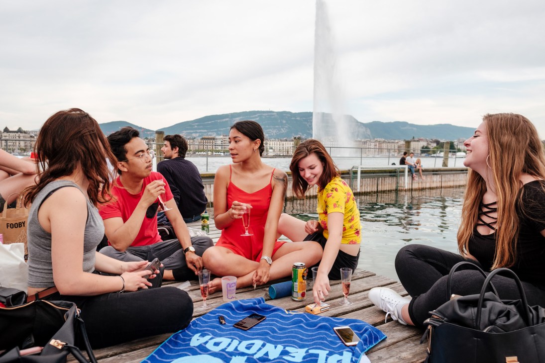 Young people socializing on a wooden deck at Bains des Pâquis with Lake Geneva and the Jet d'Eau fountain in the background.