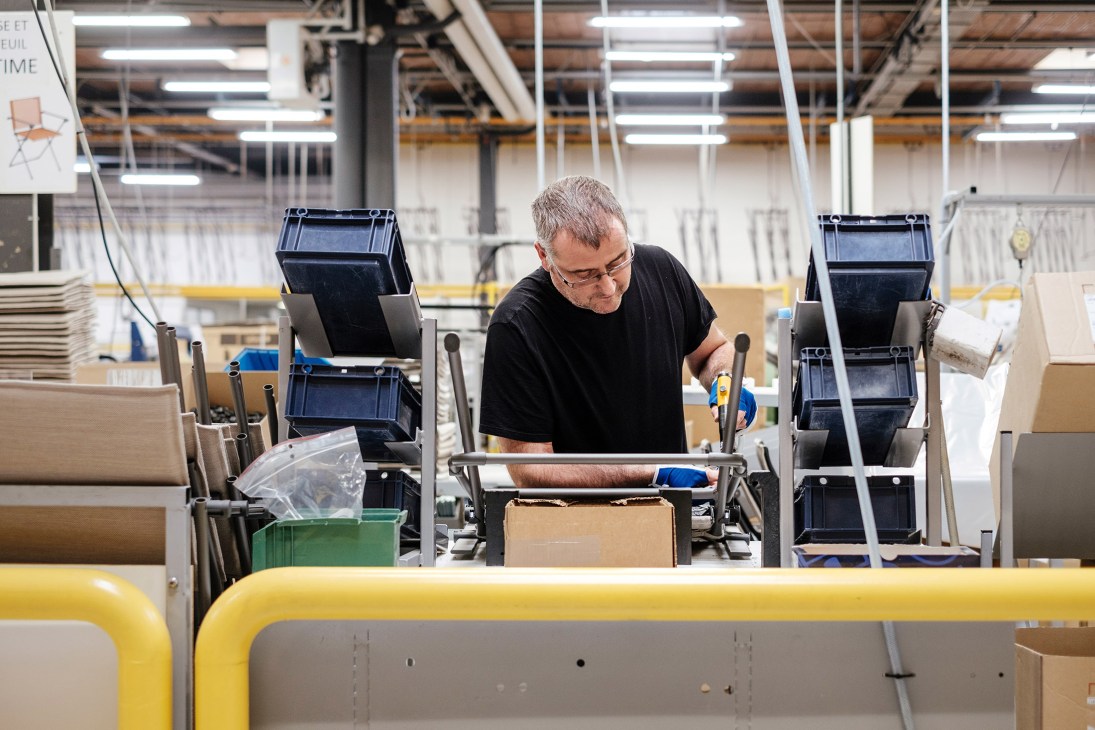 Worker assembling metal chair frames on Lafuma Mobilier production floor with storage bins and equipment visible.