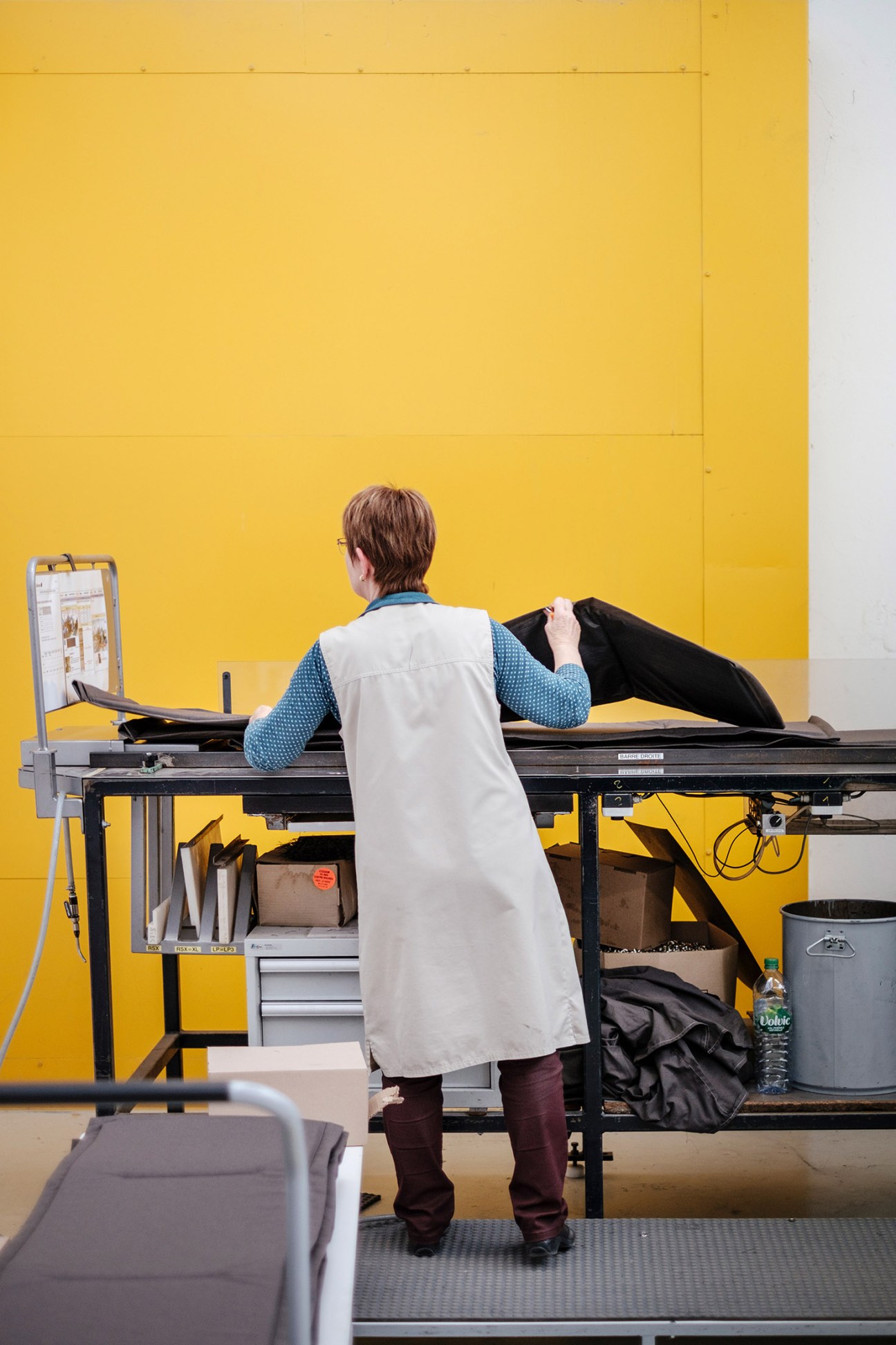 Worker in white coat testing fabric materials at a workstation against a yellow wall.