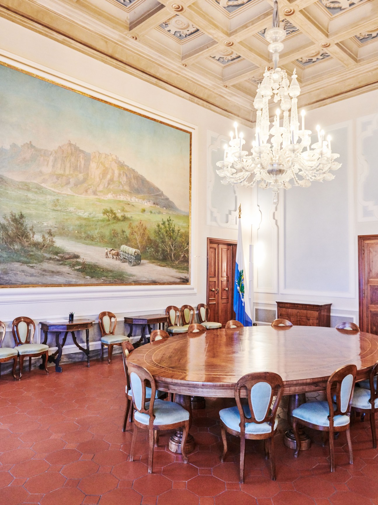 Grand meeting room with oval wooden table, ornate chandelier, and historical landscape painting of San Marino.