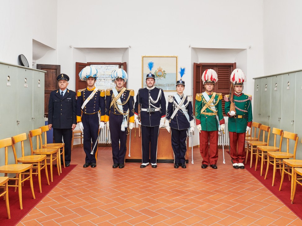 Seven Sammarinese military volunteers in ceremonial uniforms with plumed hats and ornate dress standing in a formal palace room.