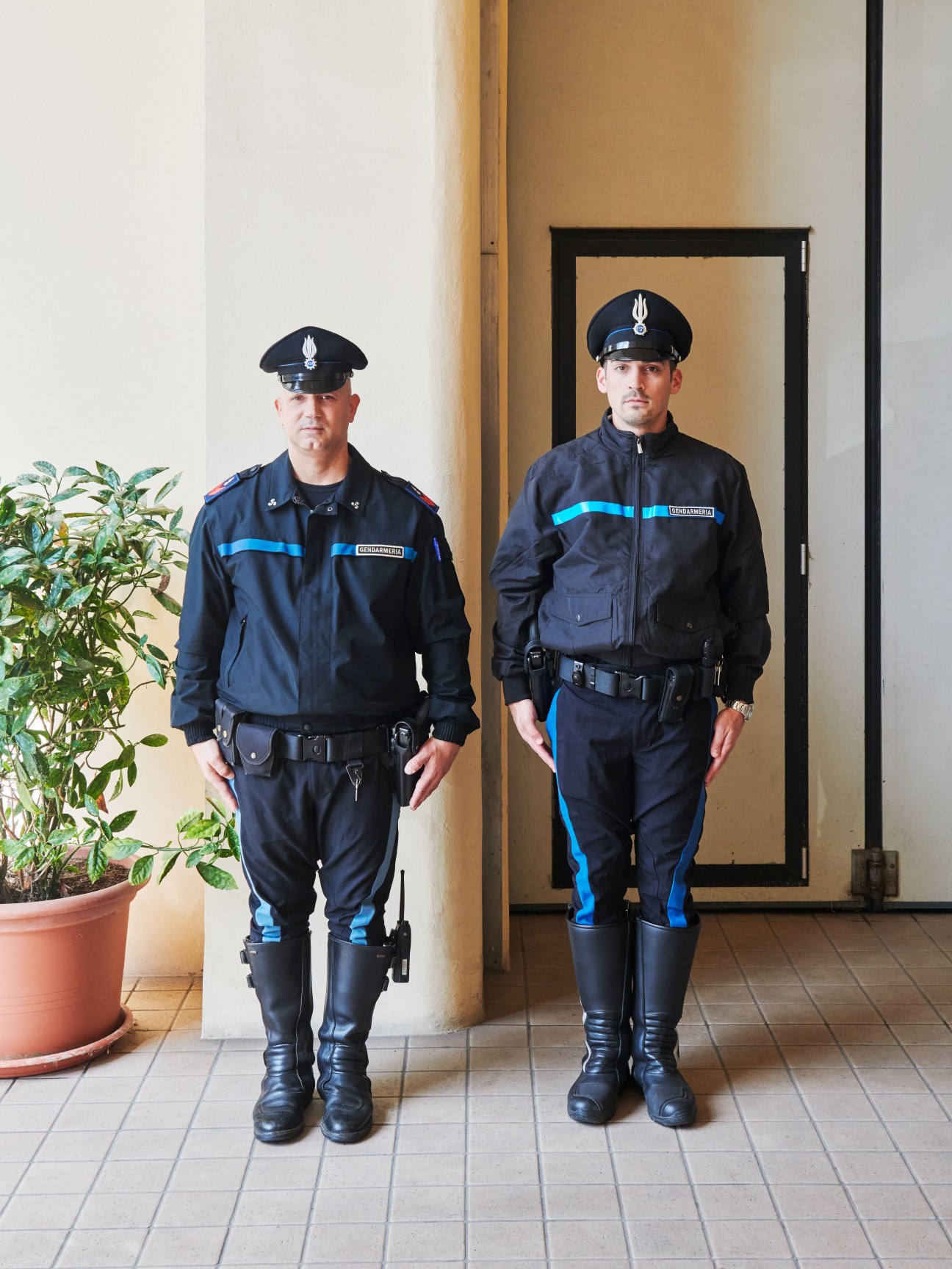 Two Gendarmerie police officers in dark uniforms with blue stripes standing in a corridor.