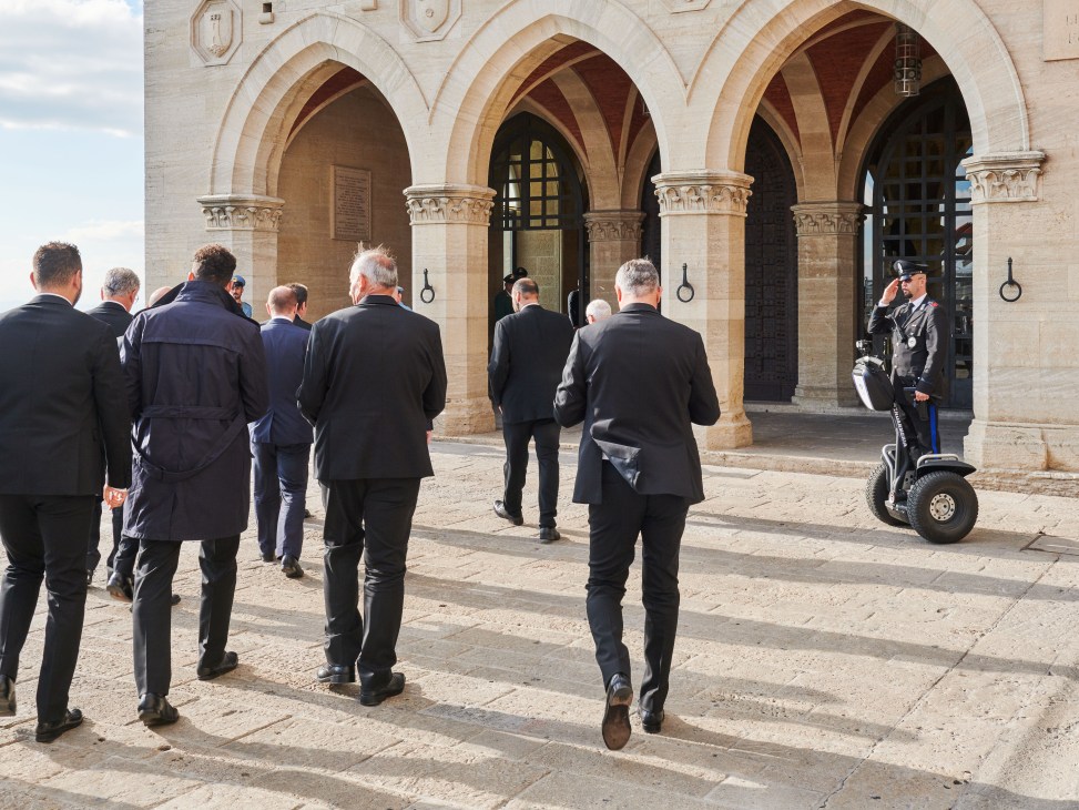 FIFA officials in dark suits arrive at the Palazzo Pubblico's Renaissance arches, with a Segway-mounted officer standing guard.