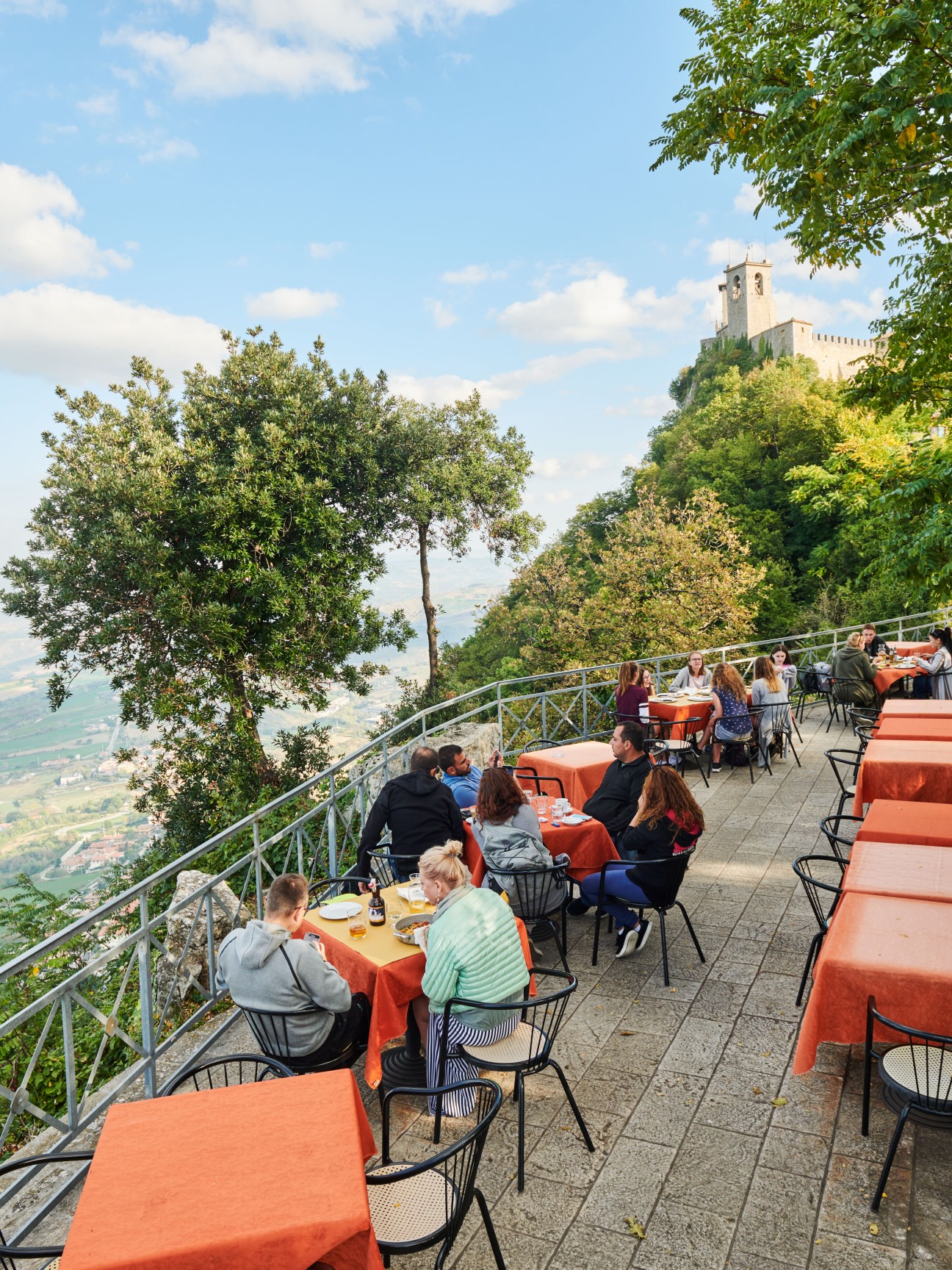 Outdoor café terrace with orange tablecloths on San Marino's fortifications overlooking the Italian countryside and a medieval tower.