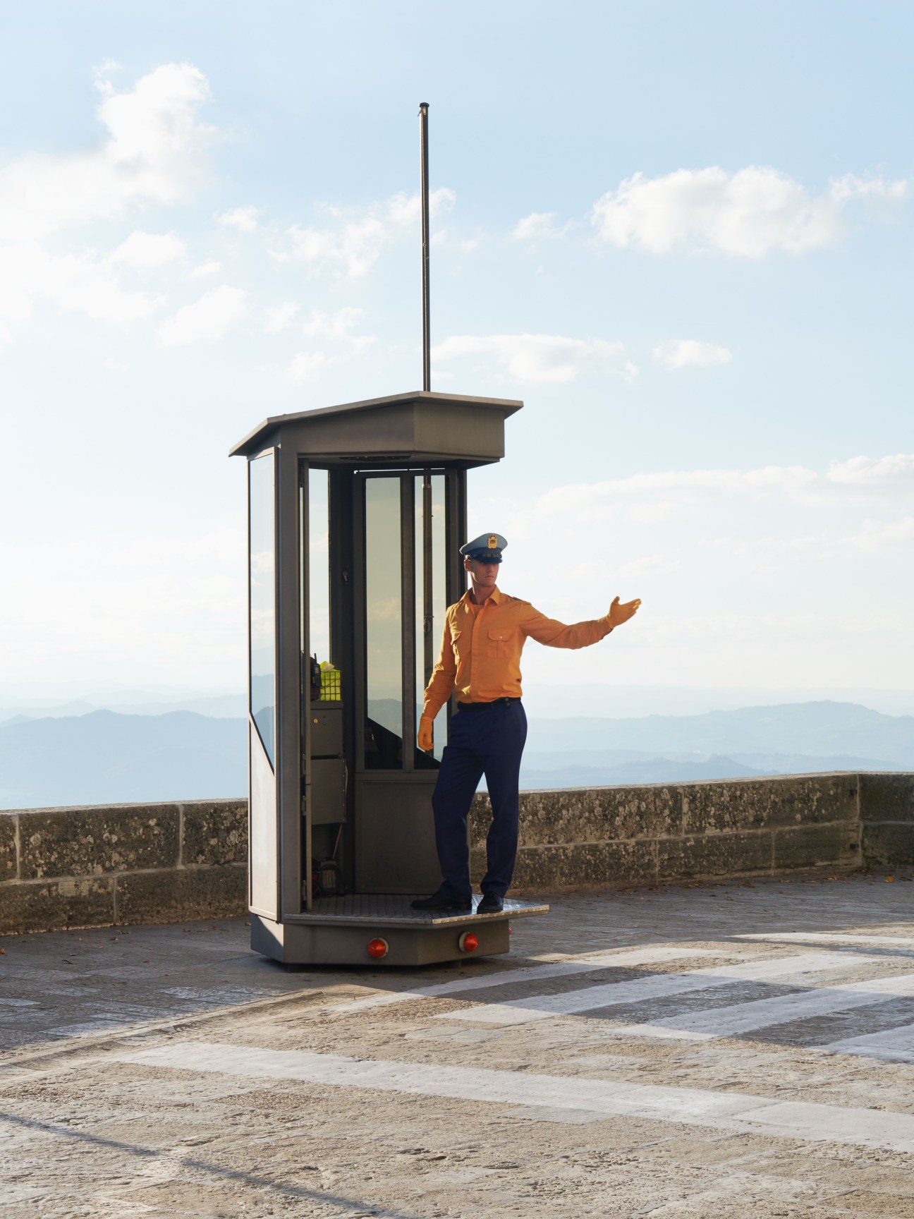 Civil police officer in orange jacket standing outside a guard booth on San Marino's fortifications with mountain views behind.