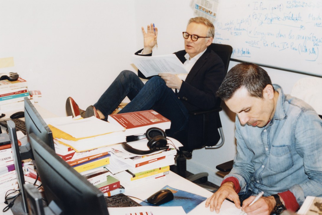 Two men in an office with stacks of books and papers, one seated reading documents while the other works at a desk.