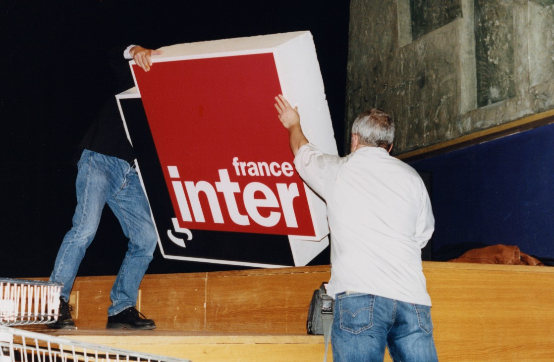 Two people holding and positioning a France Inter radio station sign on a wooden stage.