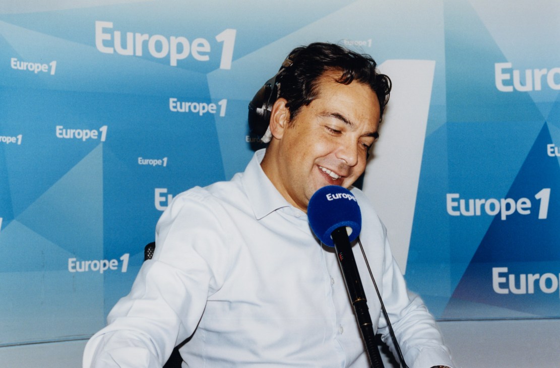 Patrick Cohen, Europe 1 morning host, smiles while speaking into a microphone at a Europe 1 branded backdrop.