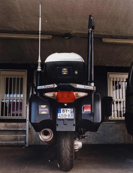 Rear view of a black motorbike with France Inter branding and radio antennas parked against a building.