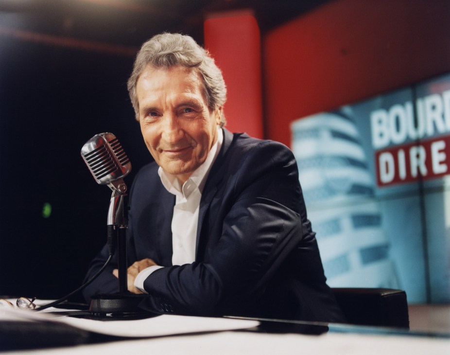 RMC host Jean-Jacques Bourdin at microphone in radio studio with red and blue branding visible behind.