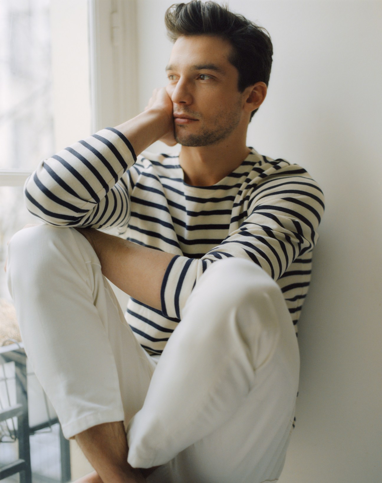 Man in striped long-sleeve shirt and white trousers sitting thoughtfully by window