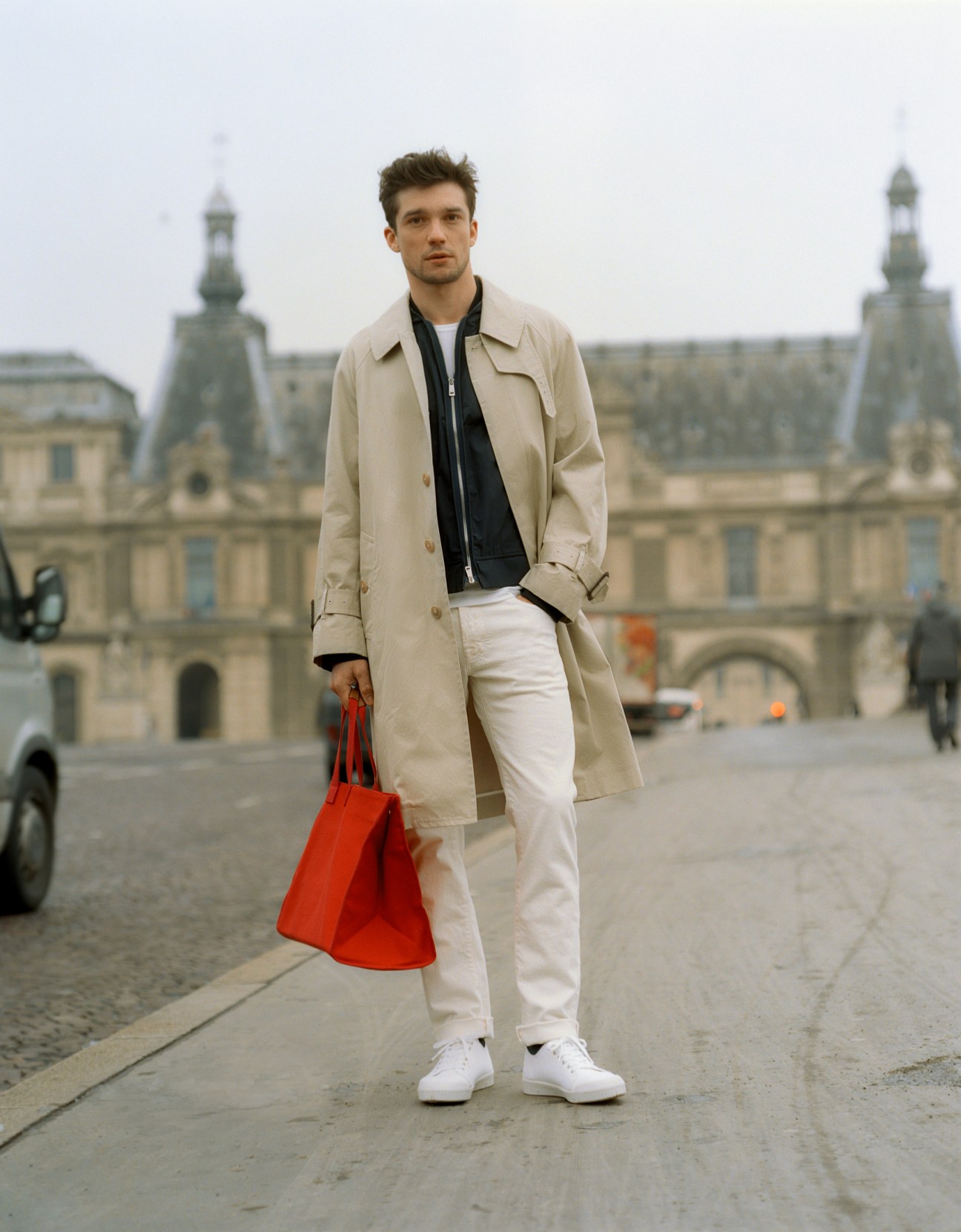 Man in beige Mackintosh coat and white jeans with red bag standing in Paris square with classical buildings.