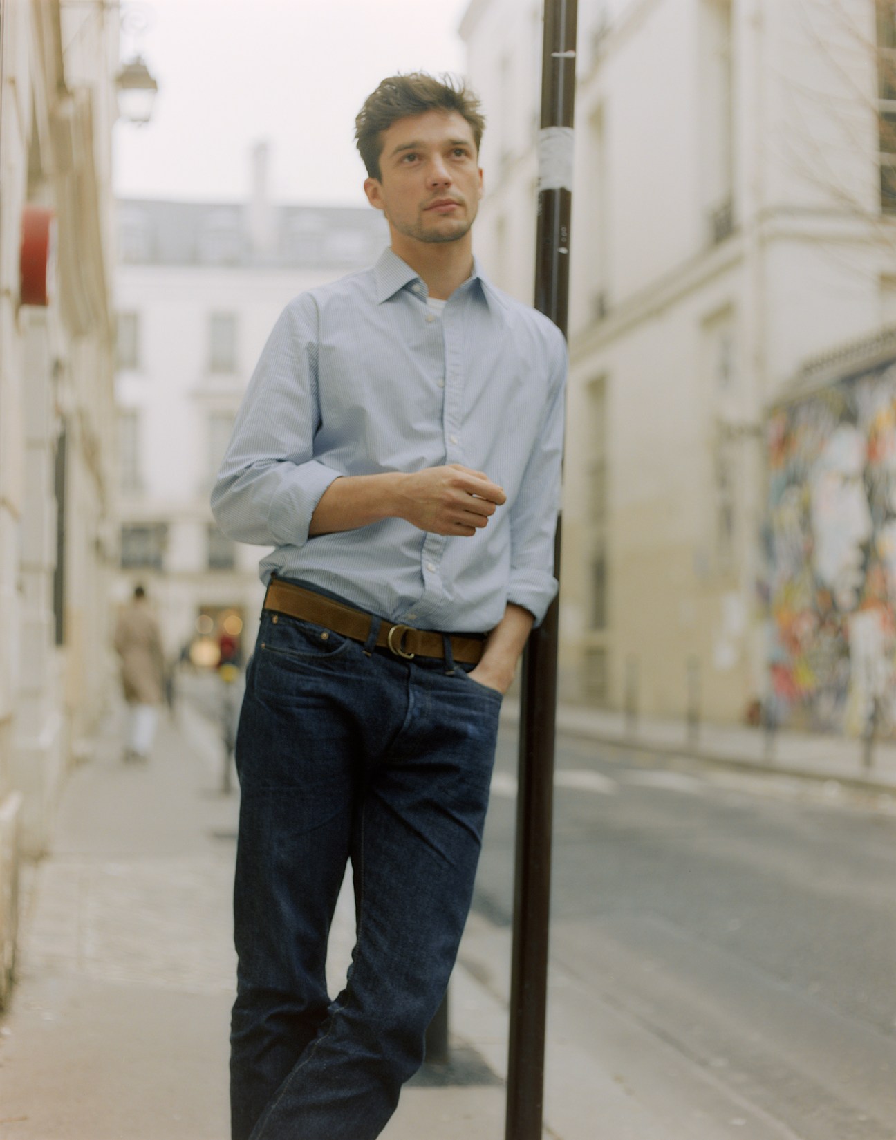 Man in light blue shirt, dark jeans and brown belt standing in a Parisian alleyway.