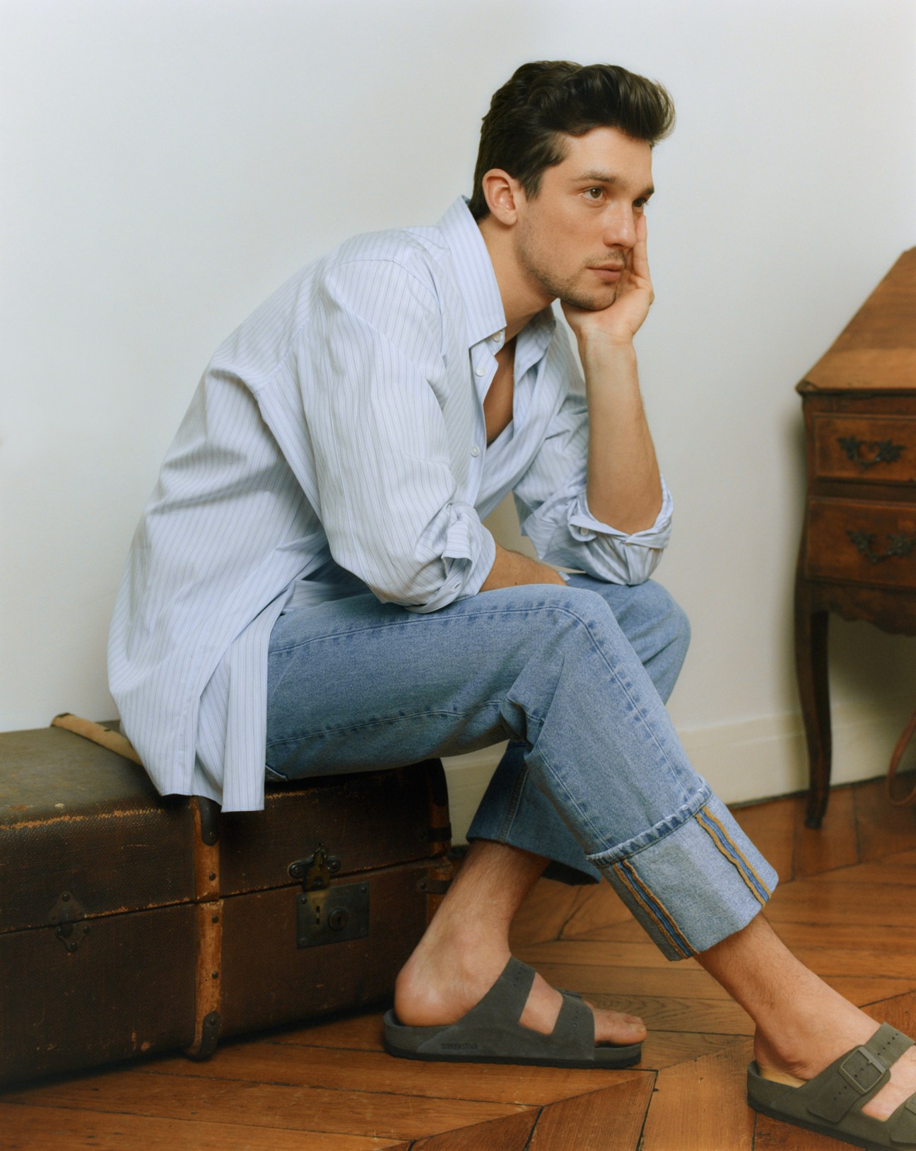 Man sitting on vintage trunk wearing light blue pinstriped shirt and jeans by Valentino with Birkenstock sandals.